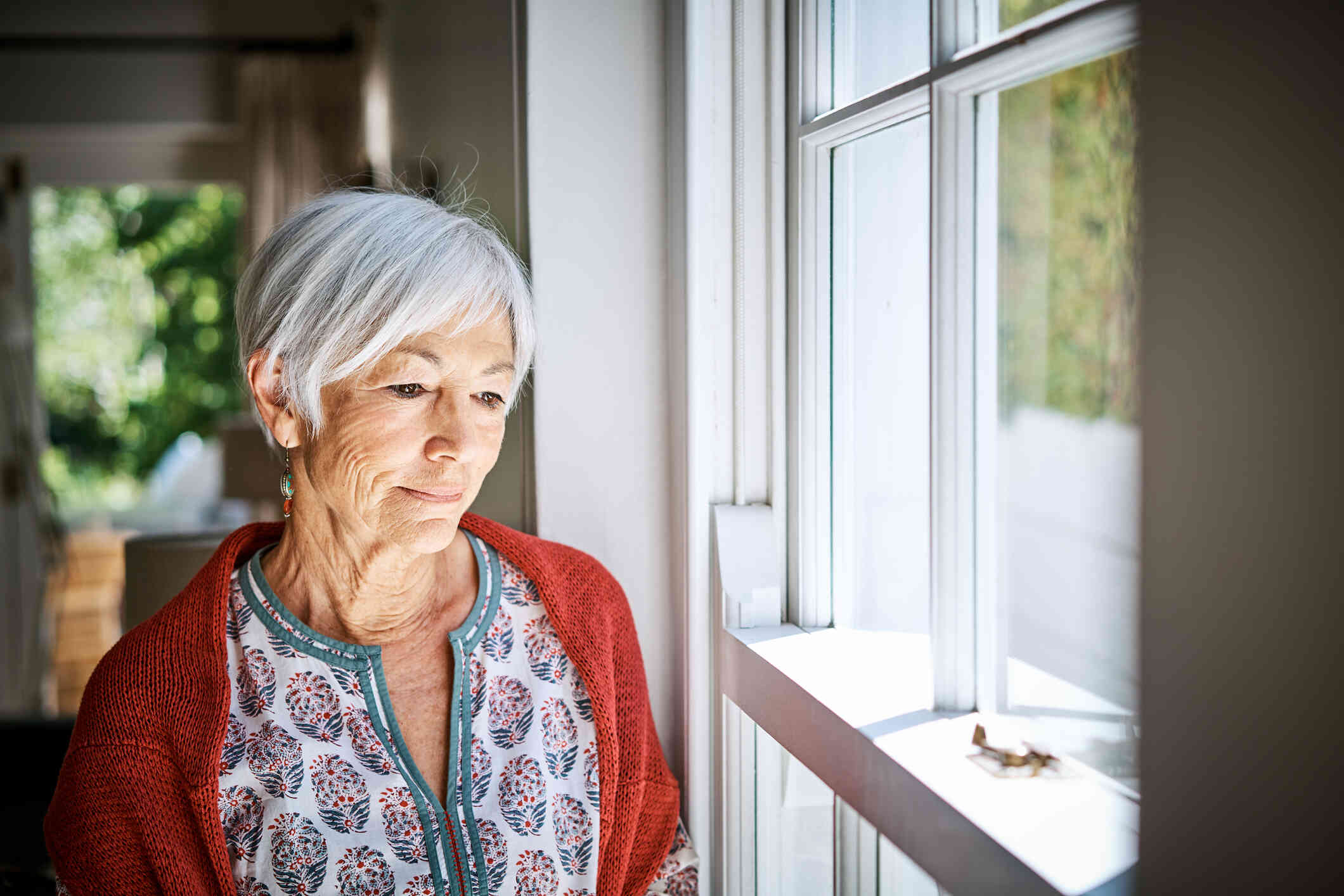 An elderly woman in a red cardigan stands in her home and gazes out of the window with a sad expression.